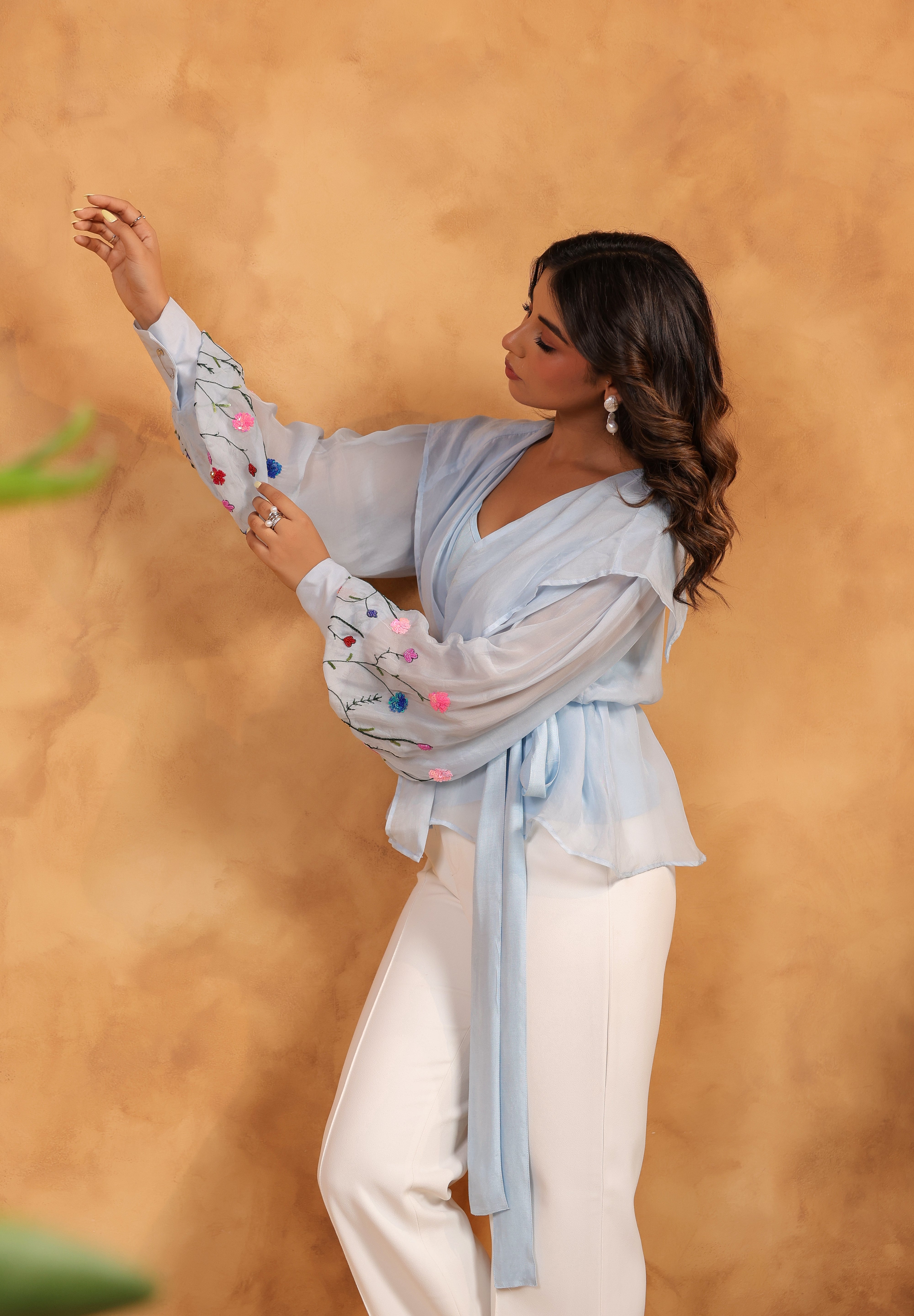 Woman wearing a light blue top with colorful pom-poms and white pants against a beige background