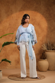 Woman in light blue top and white pants standing against a warm brown background with plants.