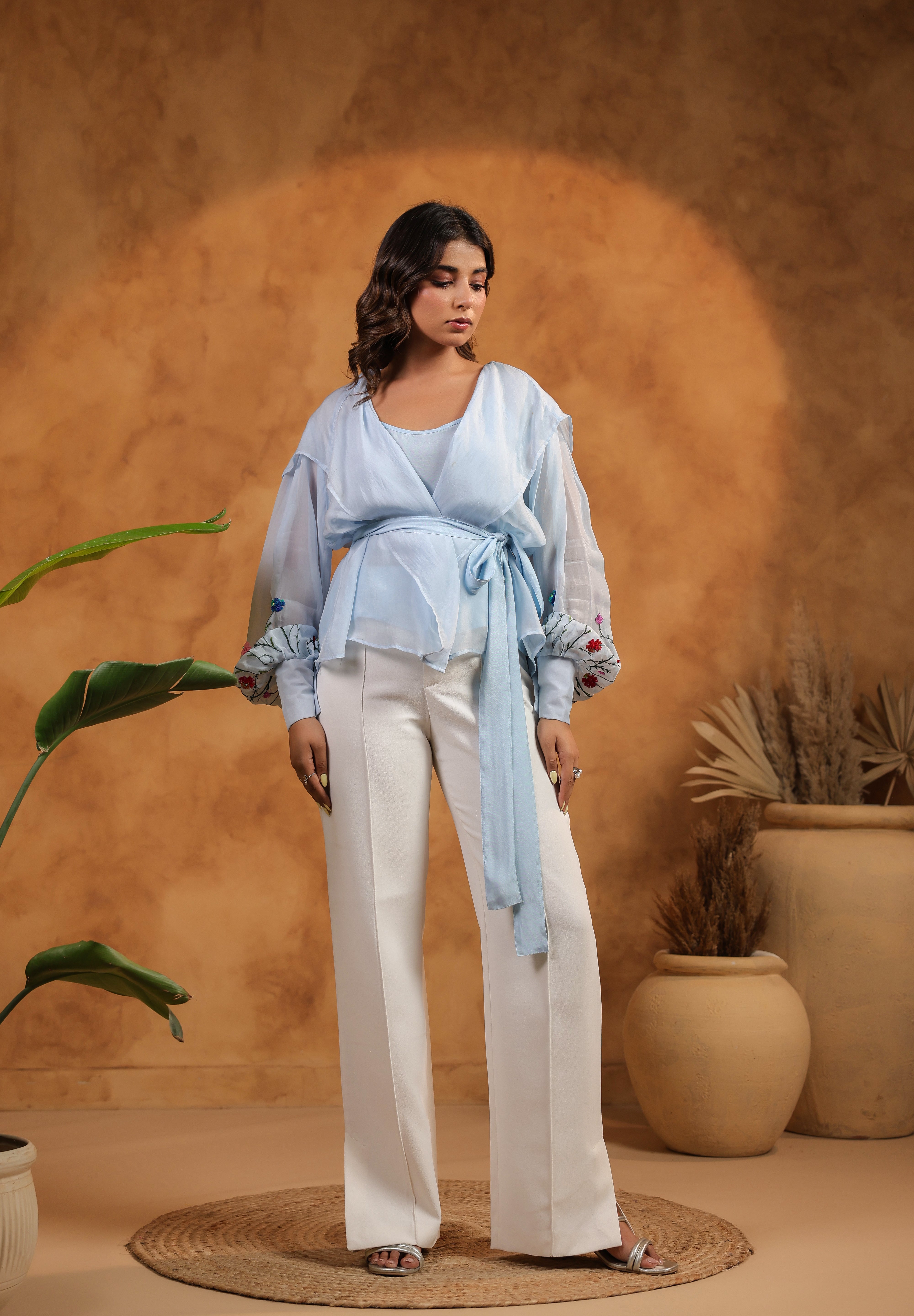 Woman in light blue top and white pants standing against a warm brown background with plants.
