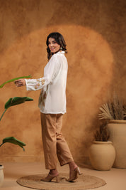Woman in a white blouse and brown pants standing against a warm-toned wall with plants.