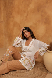 Woman in a white embroidered top and beige pants sitting against a warm-toned wall.