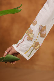 Close-up of a hand wearing a ring, holding a green leaf, with a white sleeve featuring floral embroidery on a brown background.