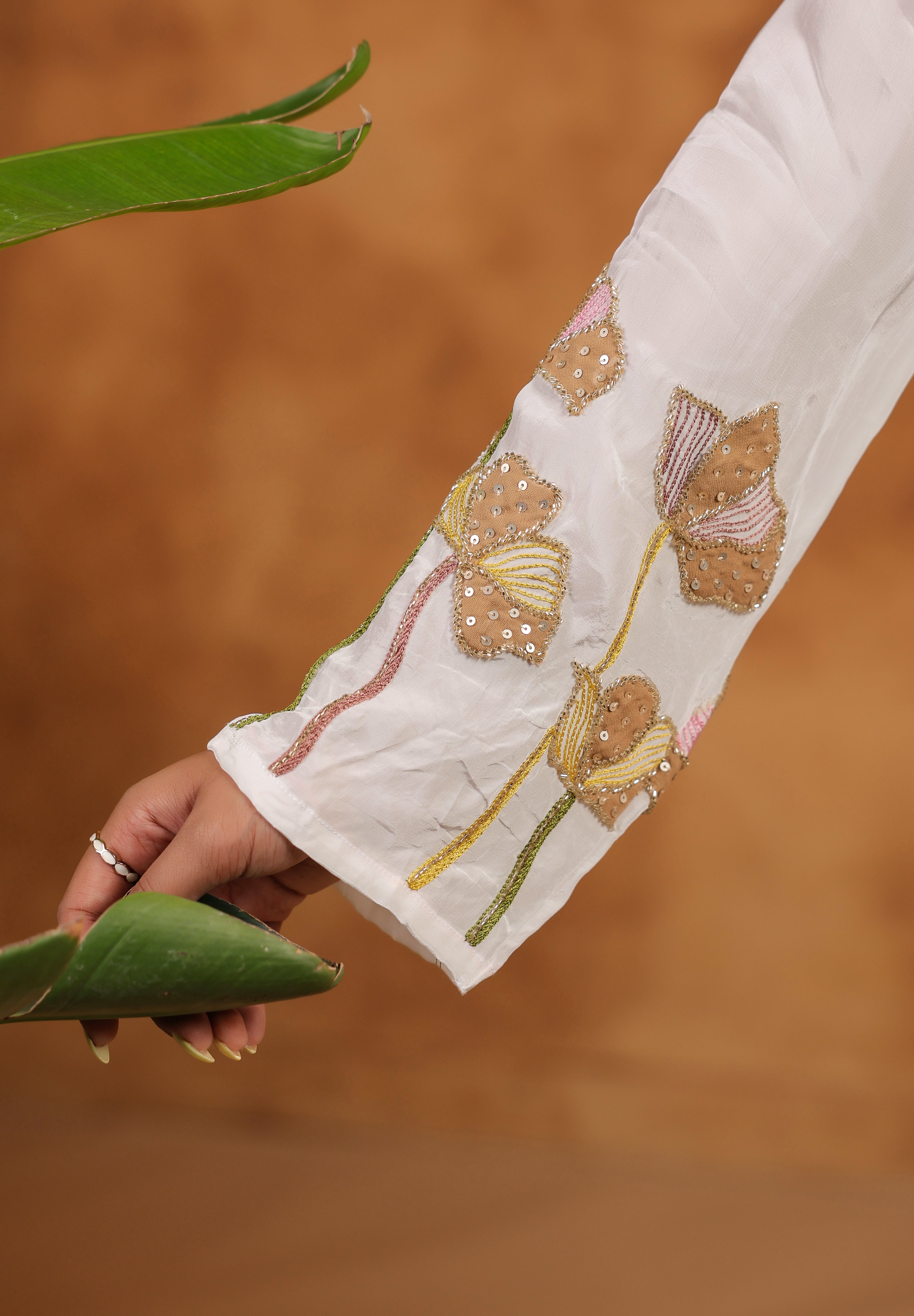 Close-up of a hand wearing a ring, holding a green leaf, with a white sleeve featuring floral embroidery on a brown background.