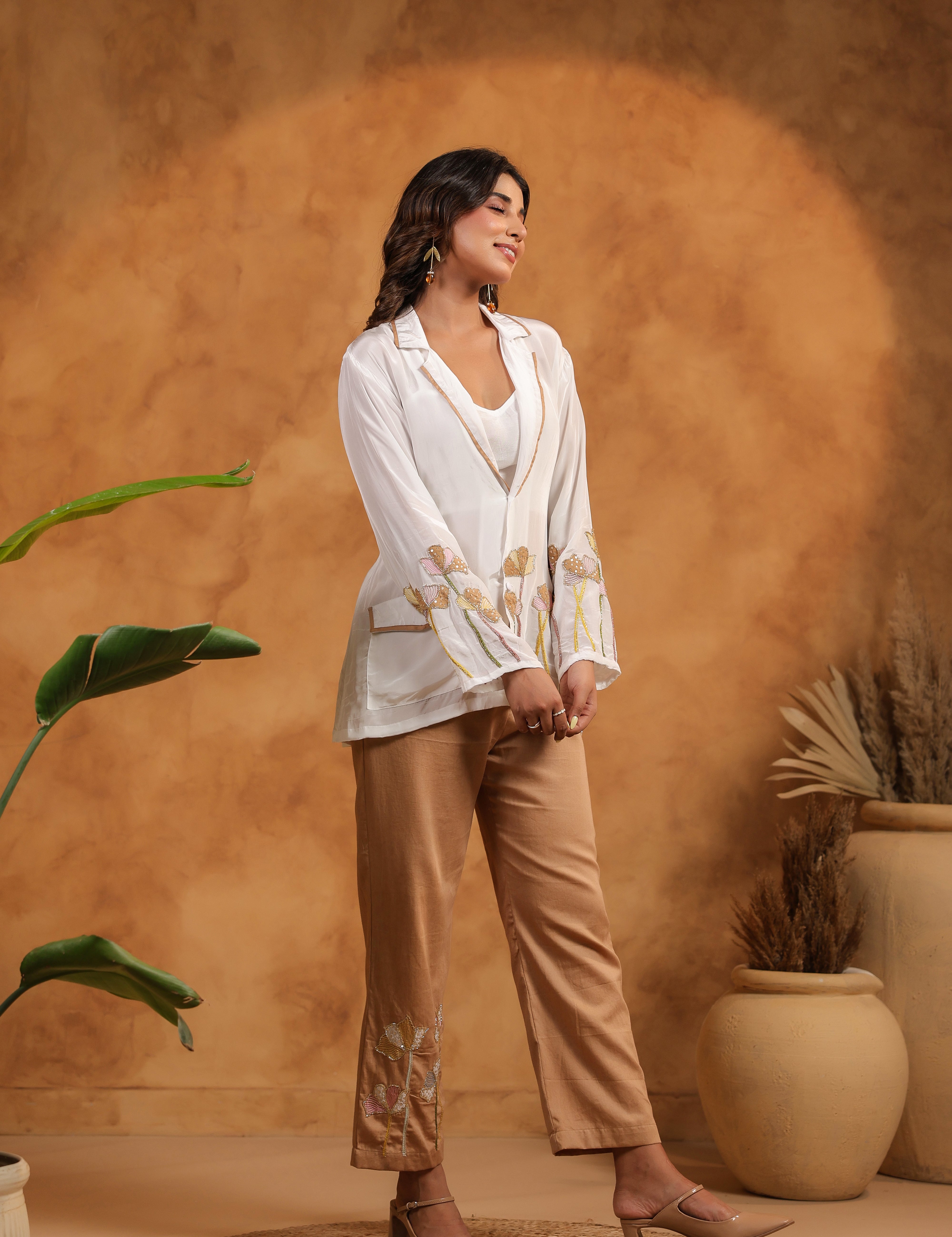 Woman in a white top and beige pants standing against a warm-toned wall with plants.