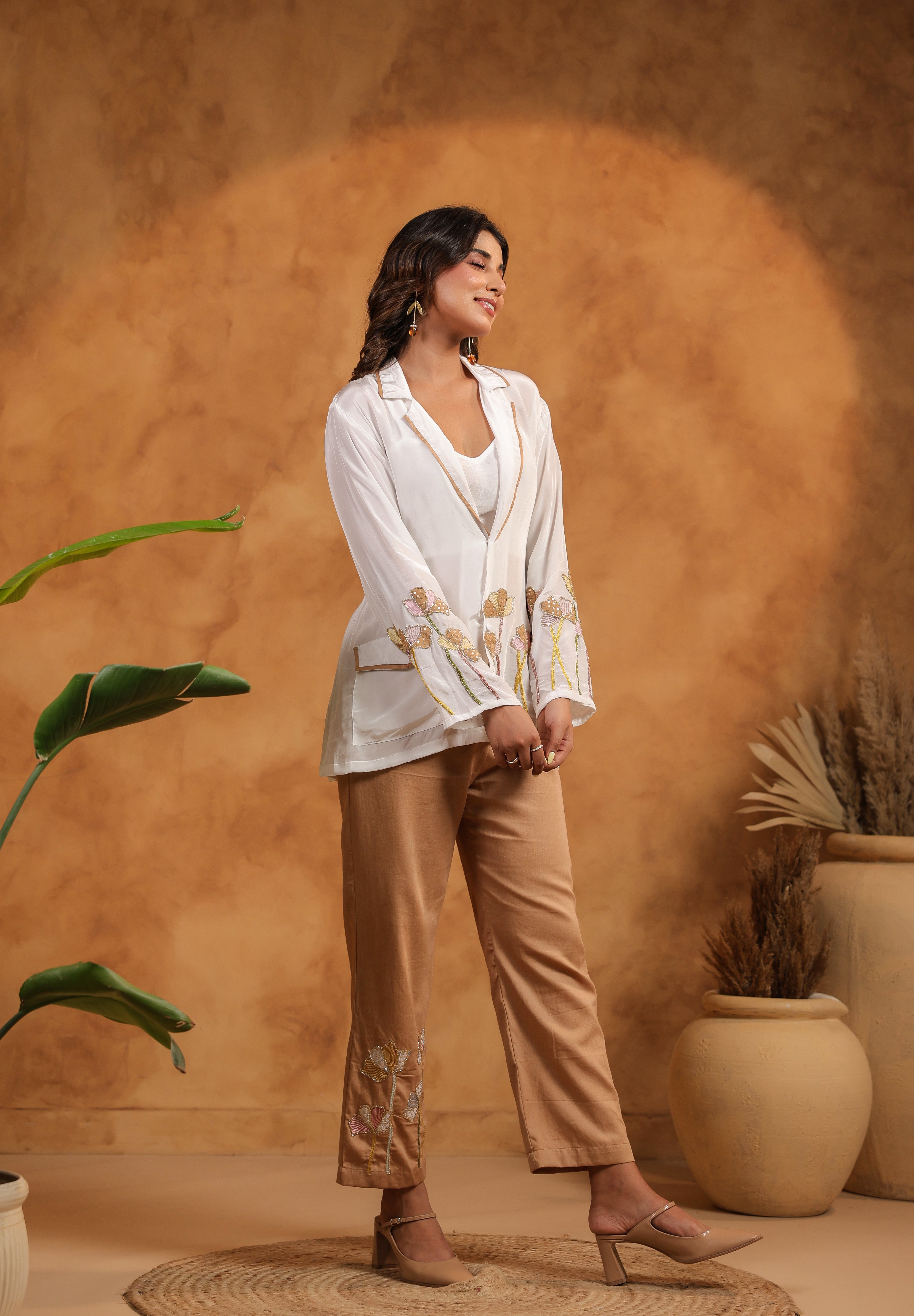 Woman in a white top and beige pants standing against a warm-toned wall with plants.