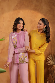 Two women in matching purple and yellow outfits standing against a warm brown background.