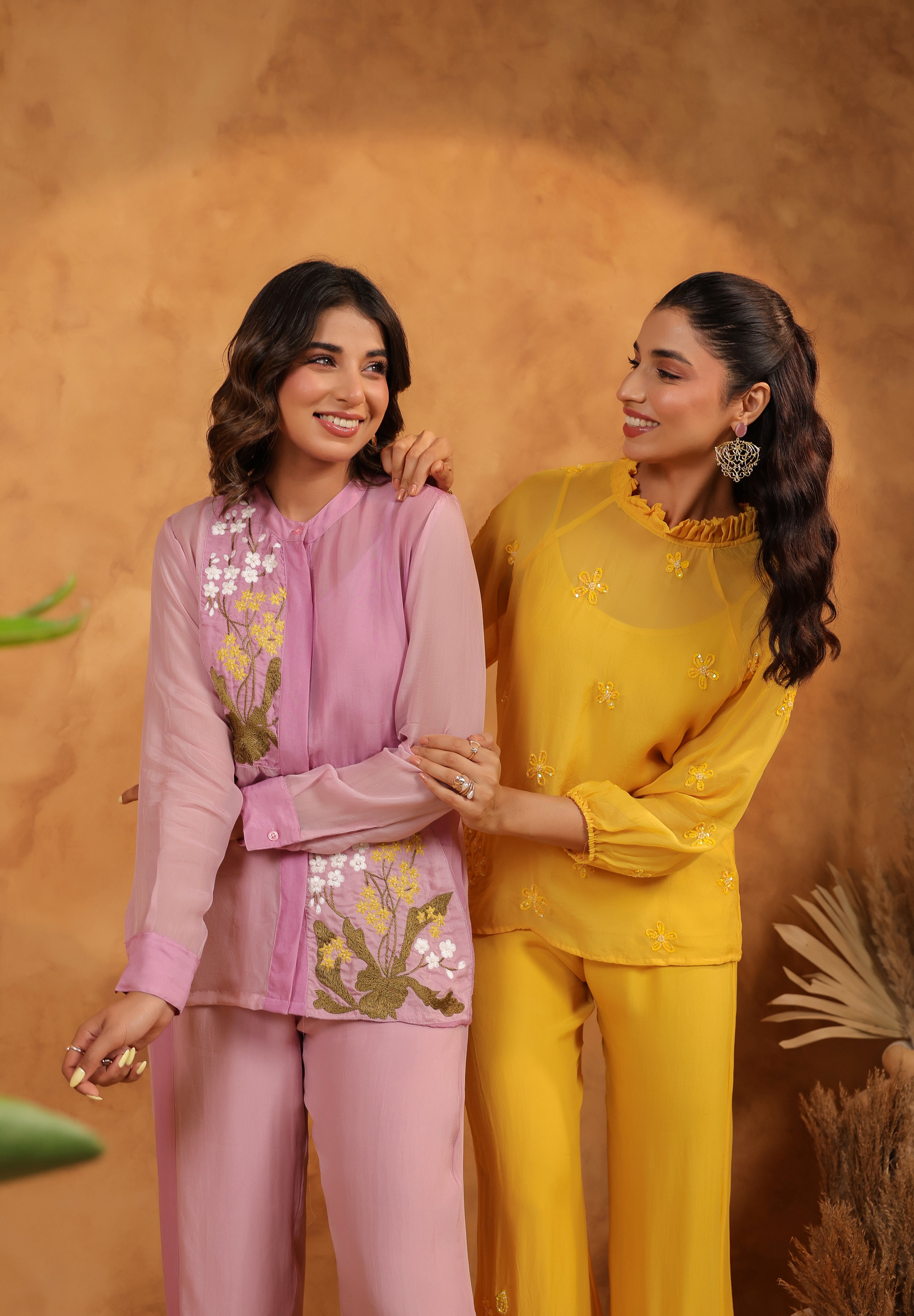 Two women in matching purple and yellow outfits standing against a warm brown background.