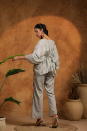 Woman in a light gray outfit standing next to a plant against a warm brown background
