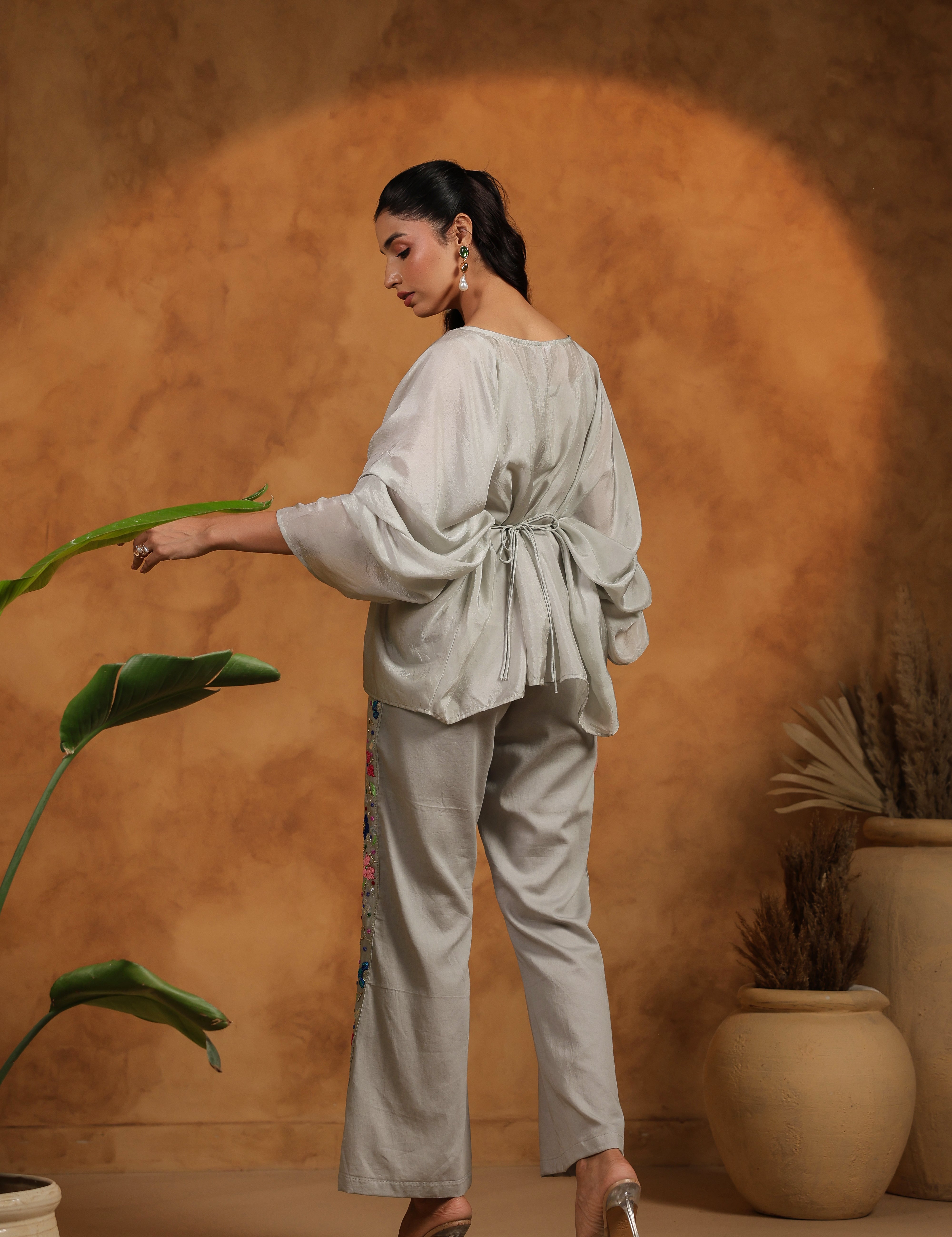Woman in a light gray outfit standing next to a plant against a warm brown background