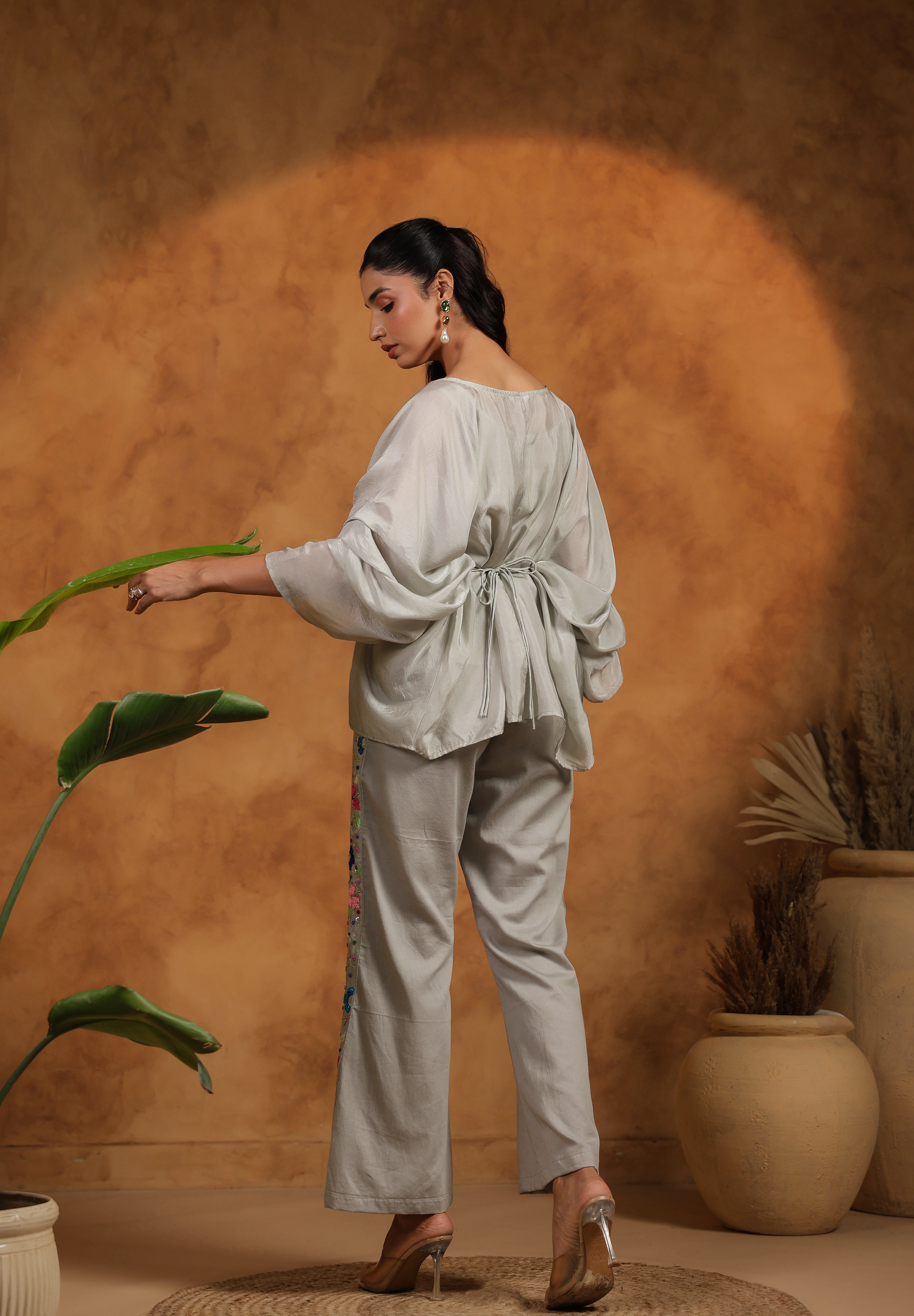 Woman in a light gray outfit standing next to a plant against a warm brown background