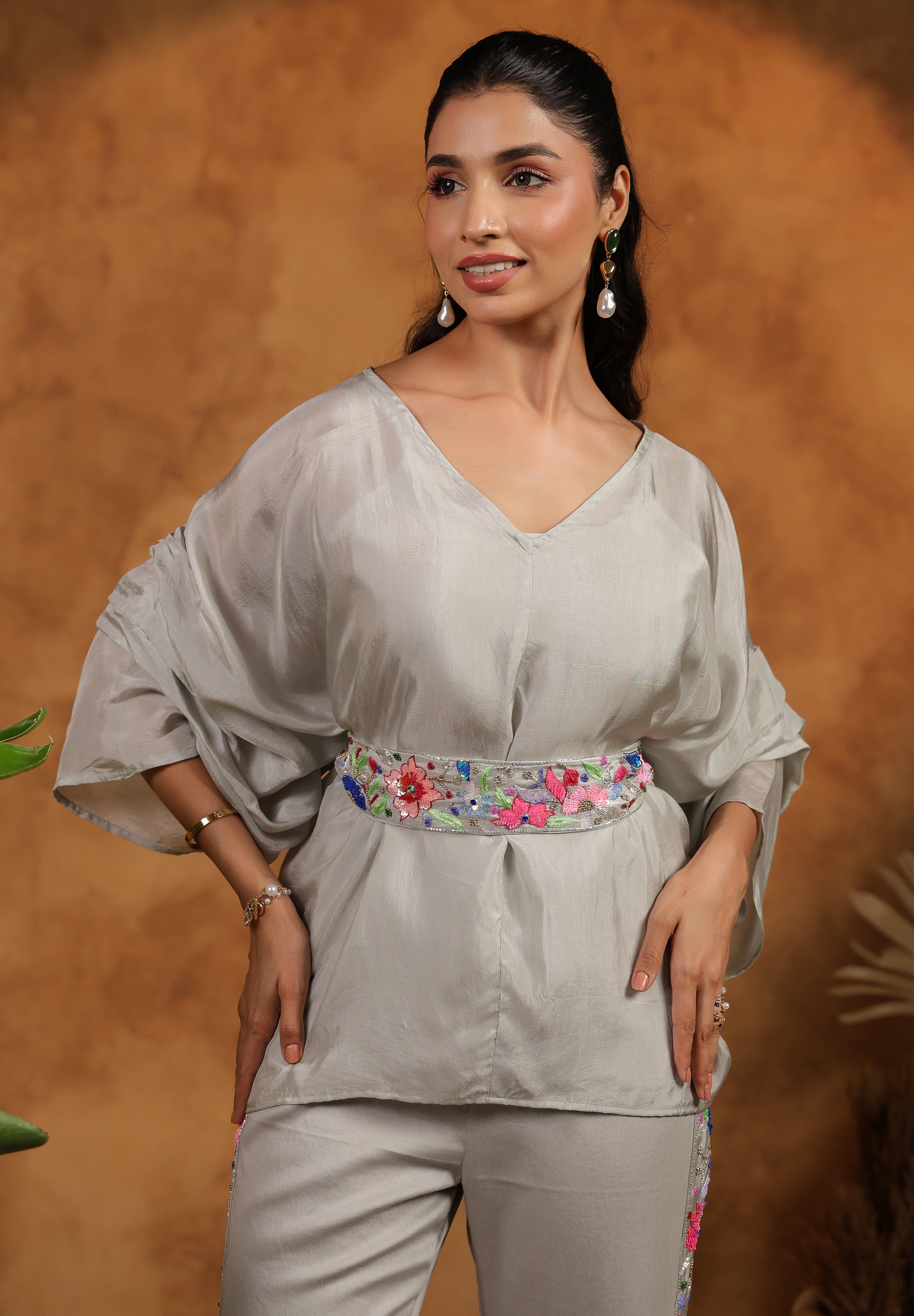 Woman wearing a light gray outfit with a floral belt against a brown background