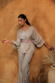 Woman in a light gray outfit with floral details against a warm-toned wall.