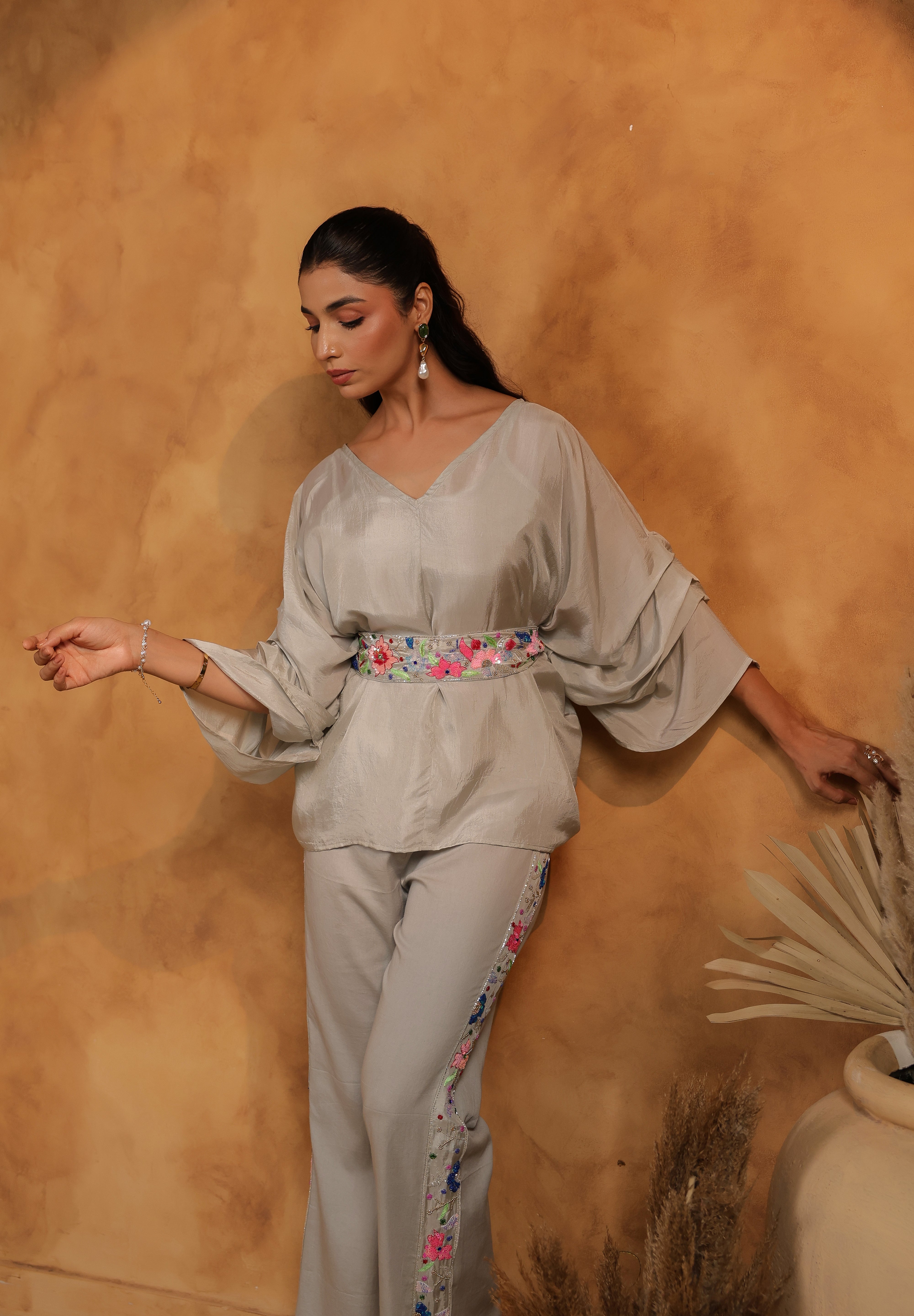 Woman in a light gray outfit with floral details against a warm-toned wall.
