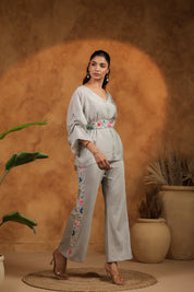 Woman in a light gray outfit with floral embroidery standing against a warm brown background.
