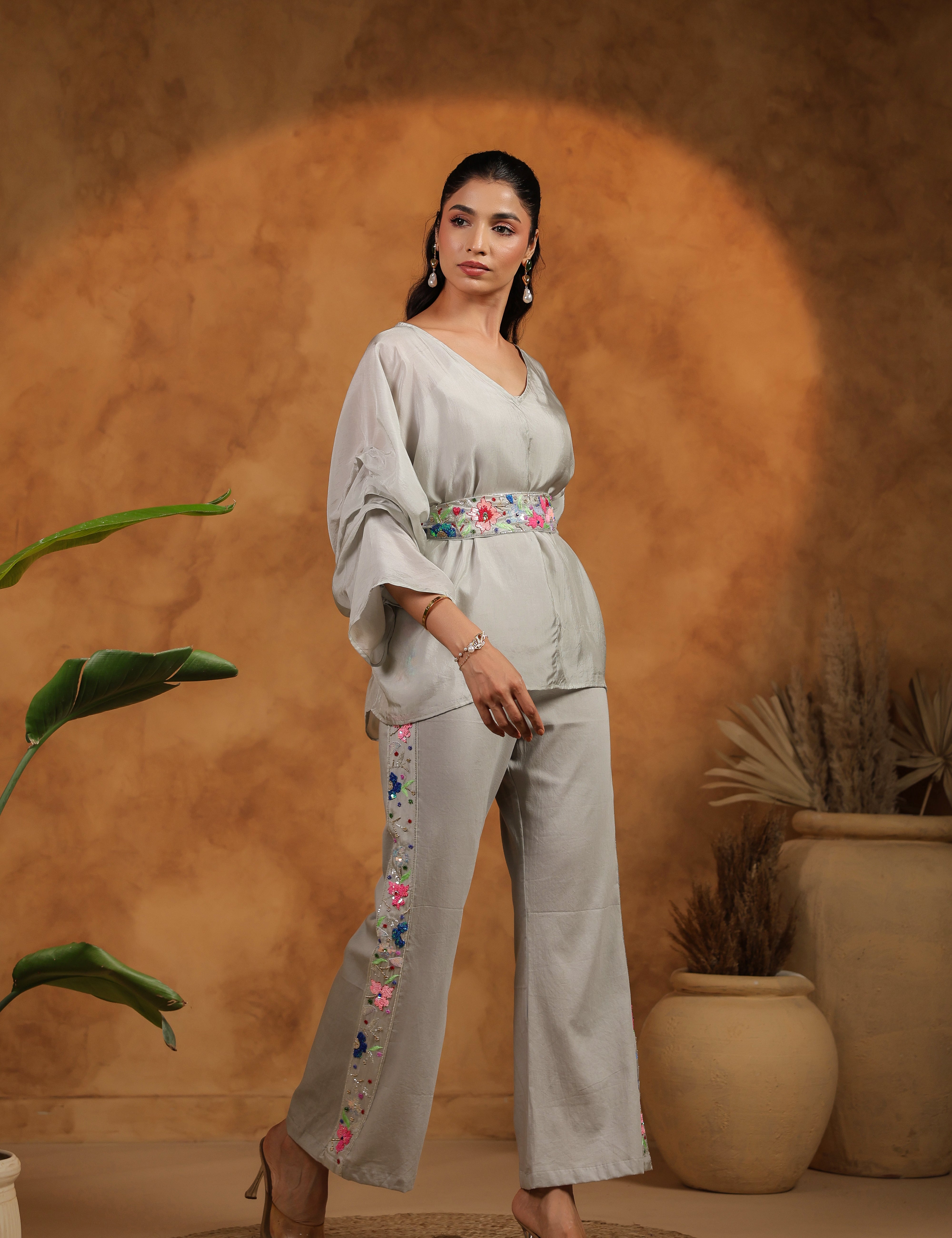 Woman in a light gray outfit with floral embroidery standing against a warm brown background.