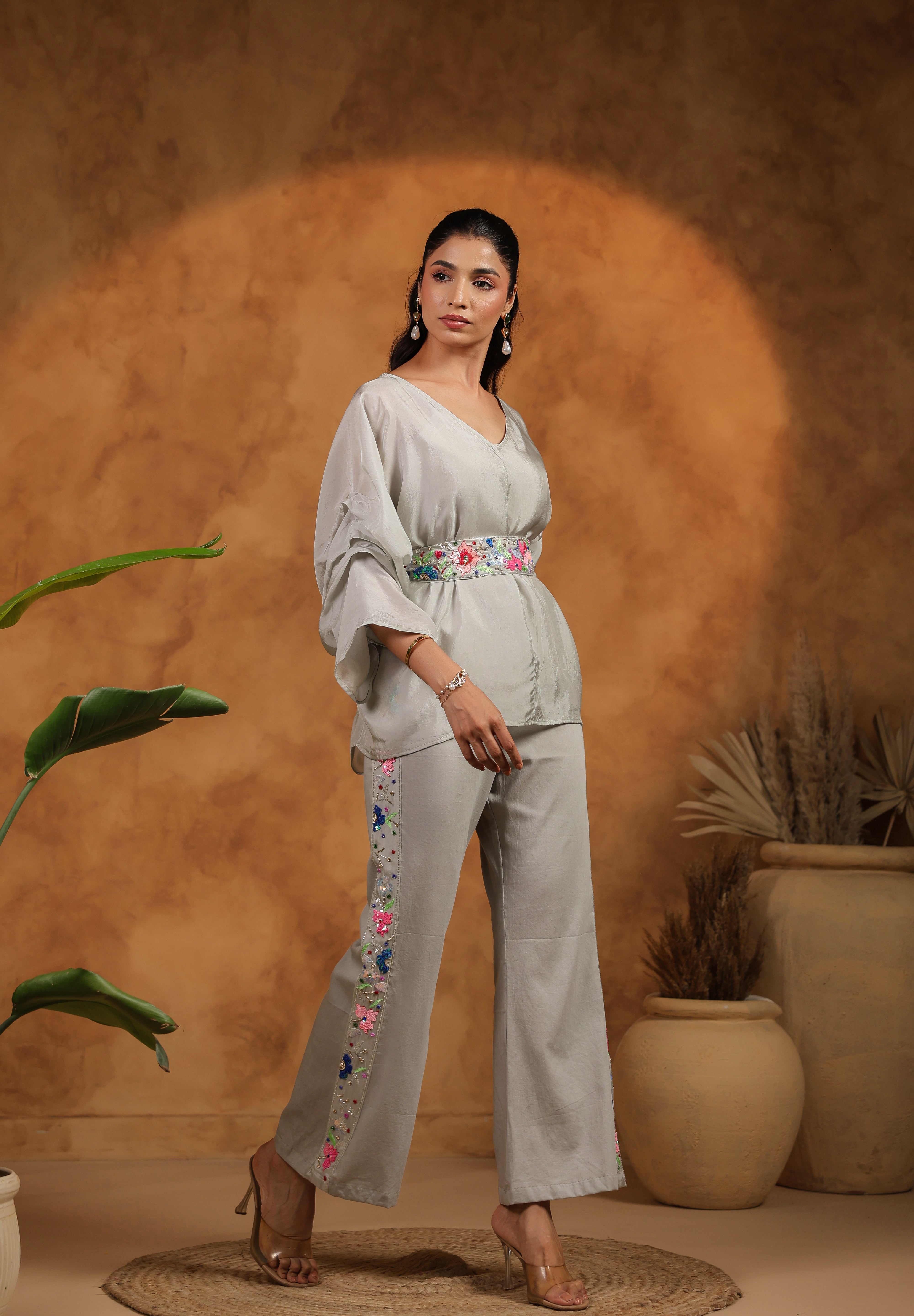 Woman in a light gray outfit with floral embroidery standing against a warm brown background.