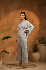 Woman wearing a light gray outfit with floral embroidery, standing against a warm-toned wall with plants.