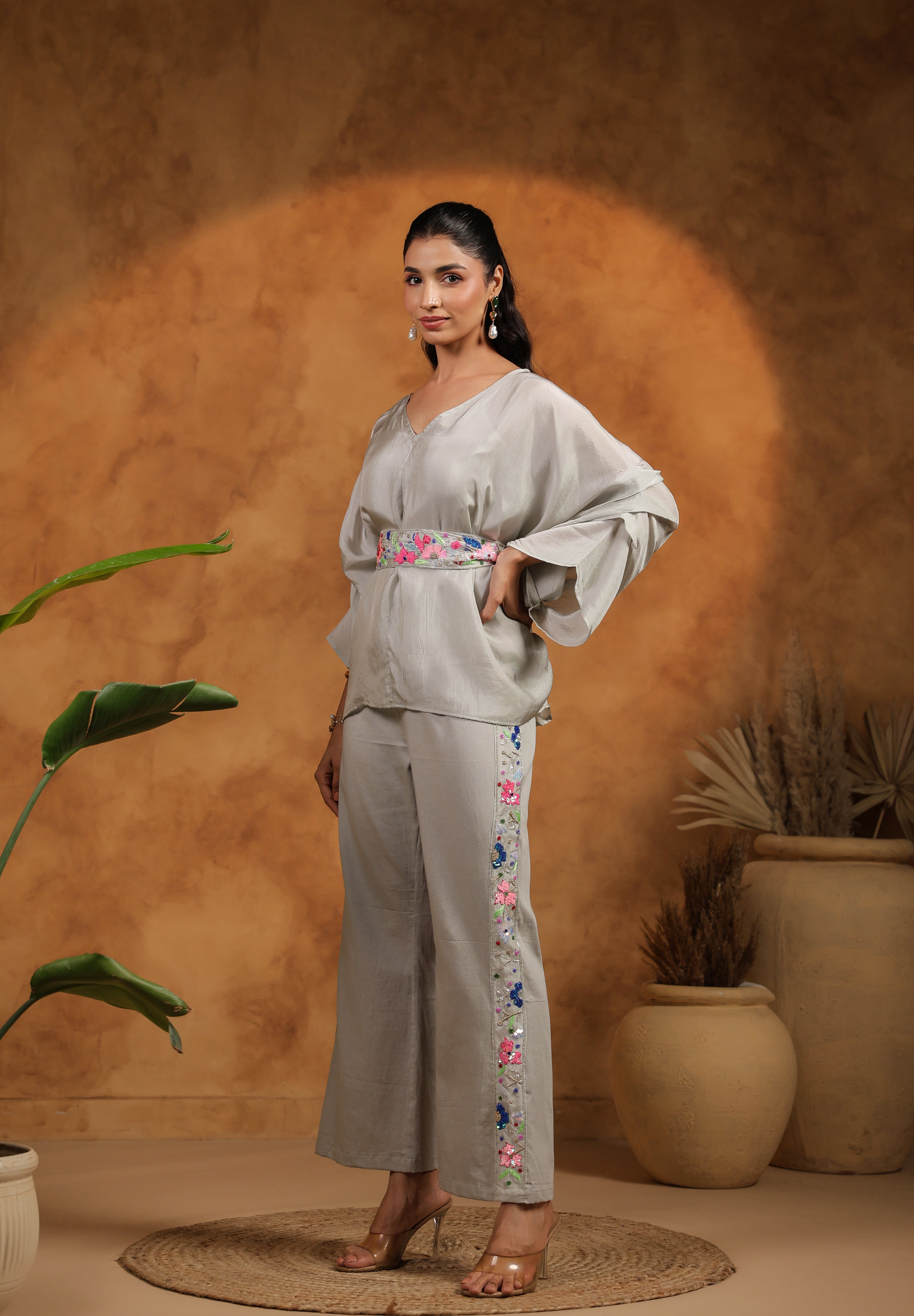 Woman wearing a light gray outfit with floral embroidery, standing against a warm-toned wall with plants.