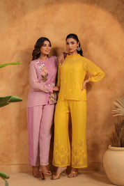 Two women in purple and yellow outfits standing against a beige wall with plants.