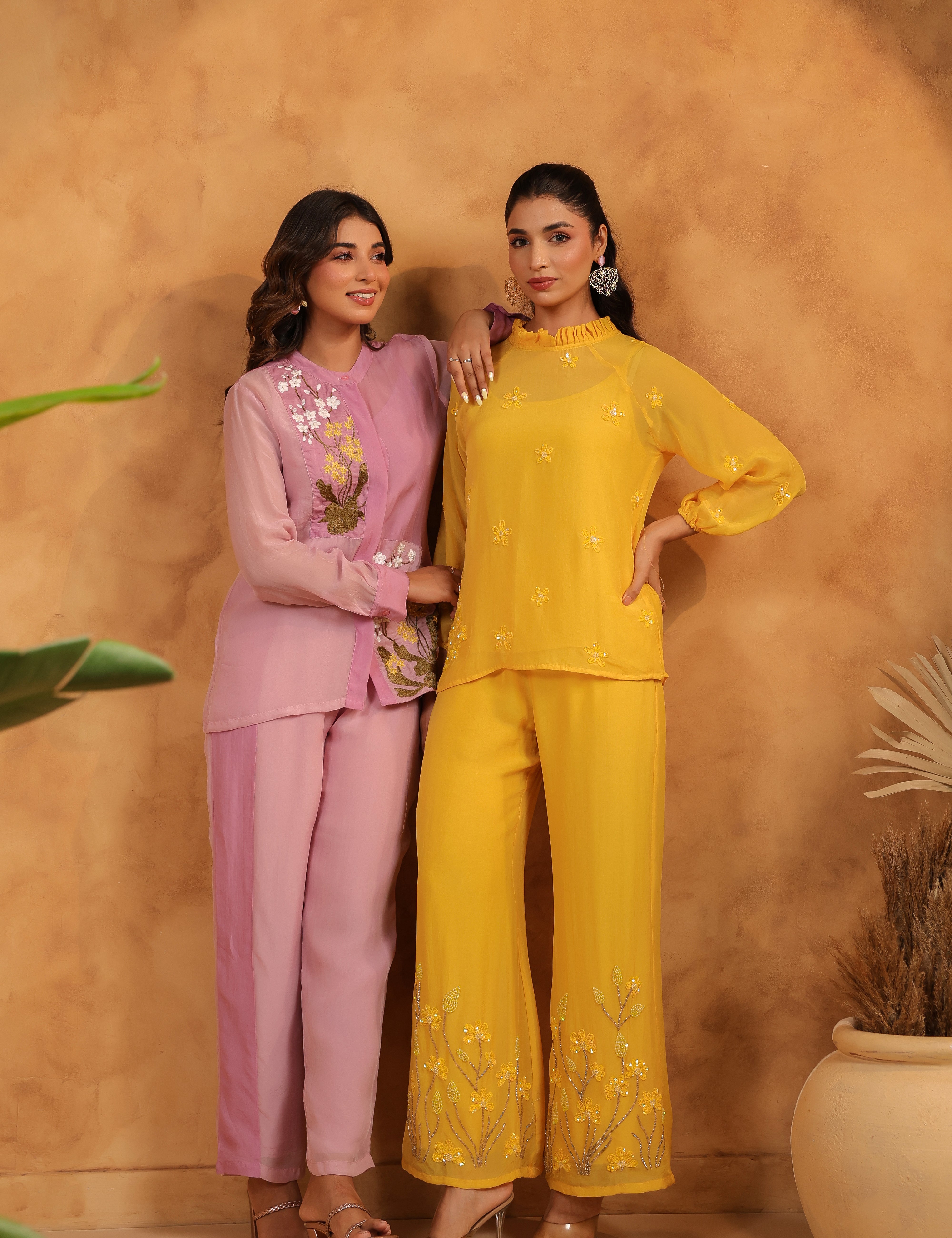 Two women in purple and yellow outfits standing against a beige wall with plants.