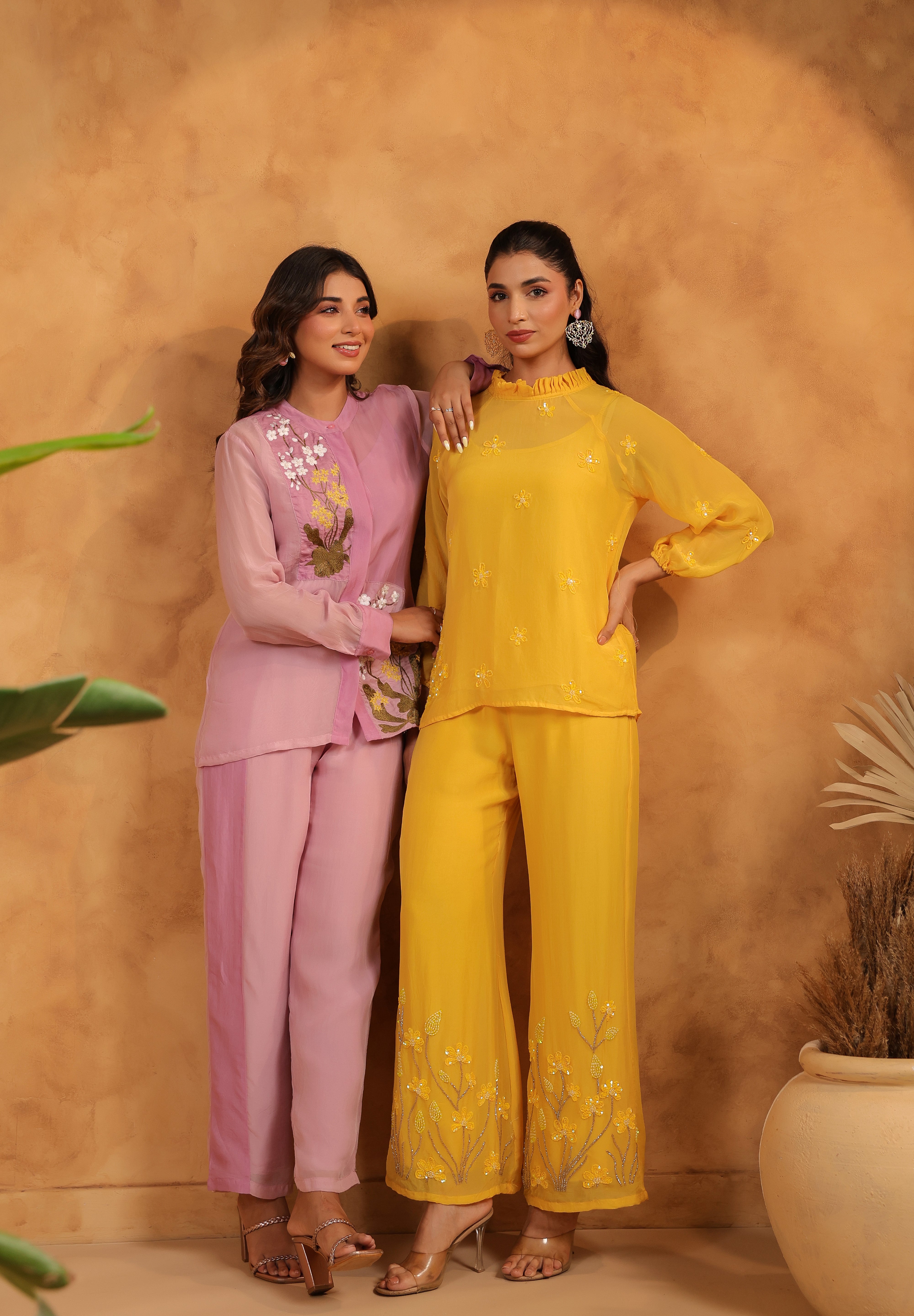 Two women in purple and yellow outfits standing against a beige wall with plants.