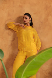 Woman in a yellow outfit standing against an orange wall with green leaves in the foreground.