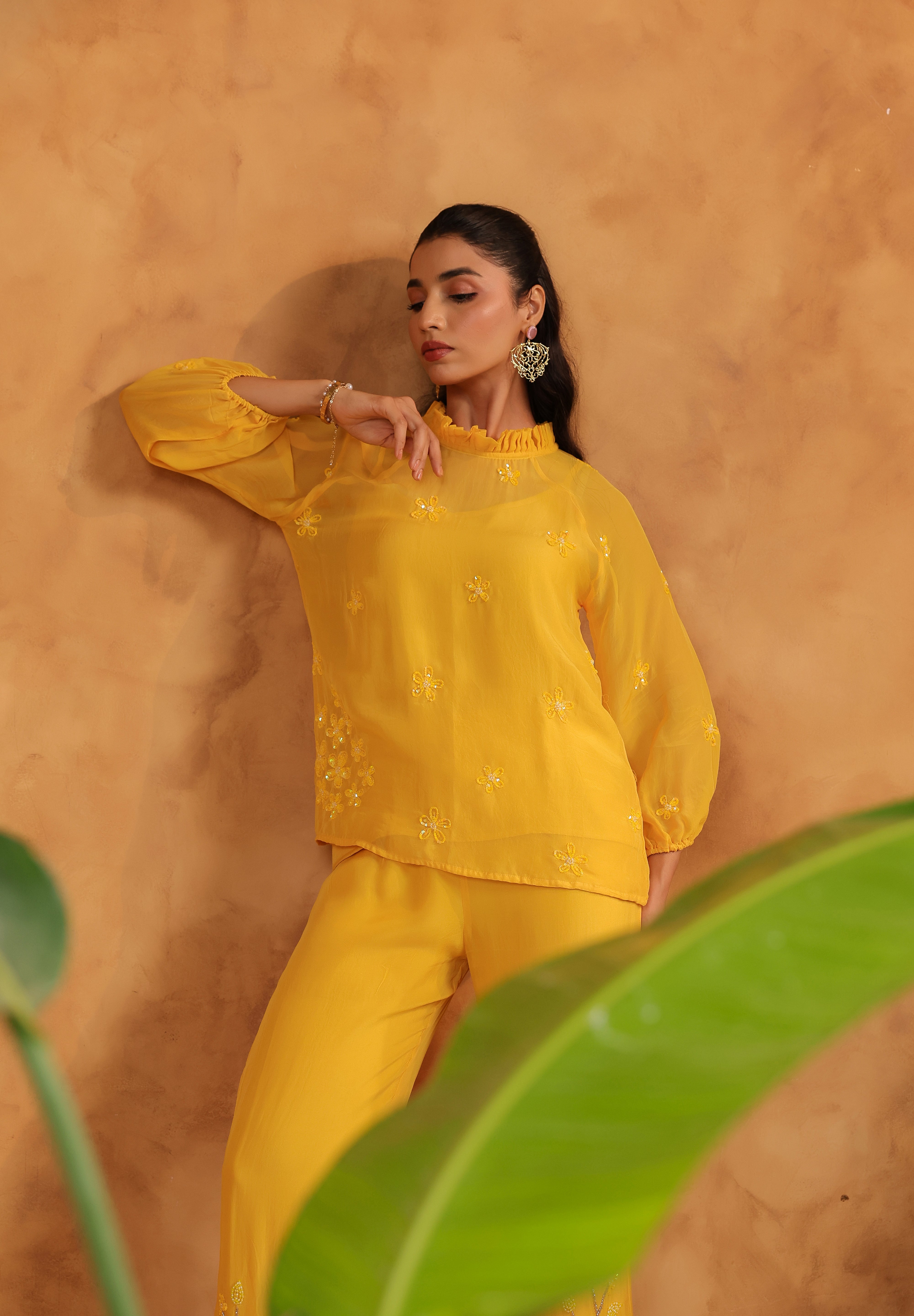 Woman in a yellow outfit standing against an orange wall with green leaves in the foreground.